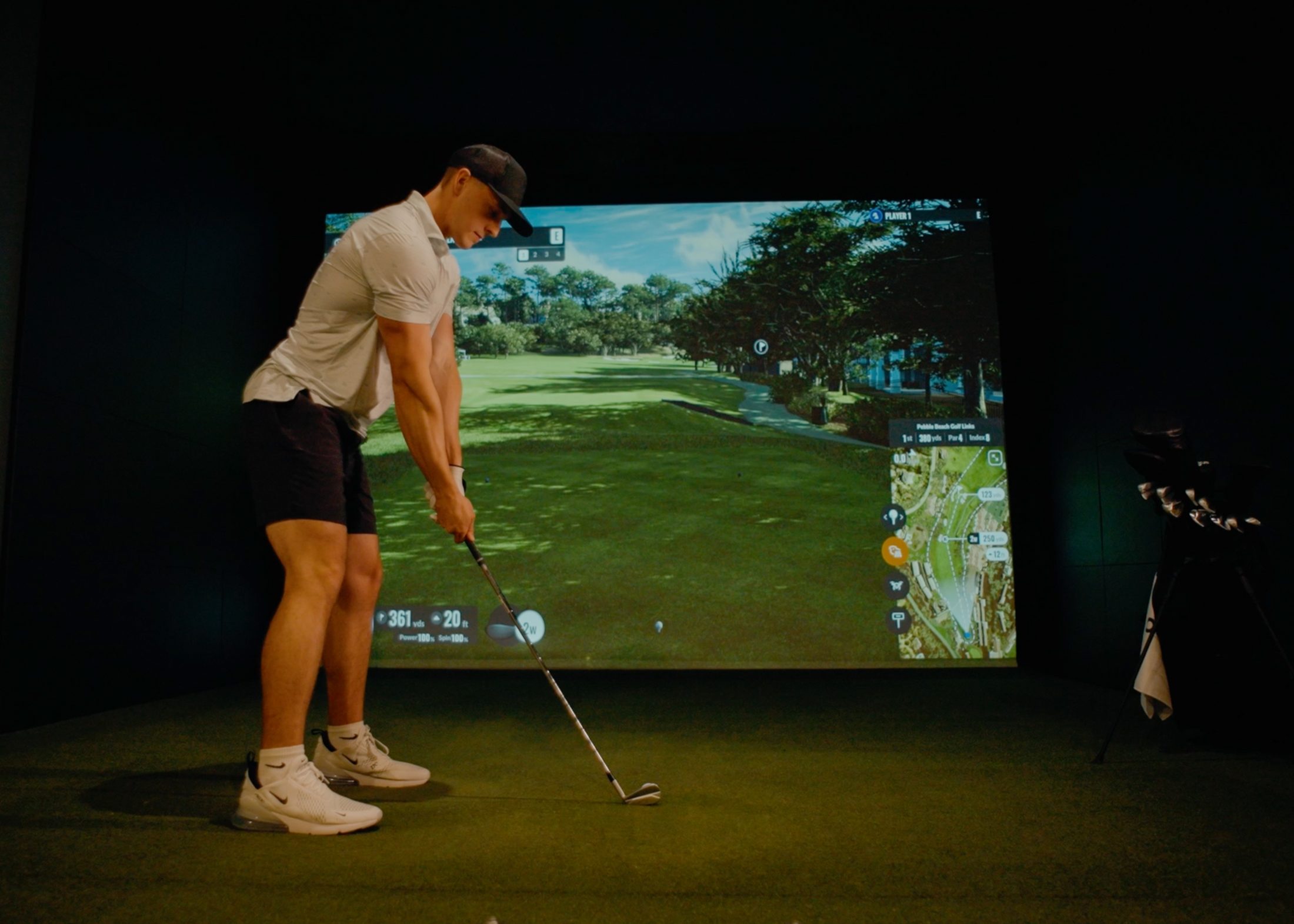 A golfer addressing the ball in a darkened simulator bay, the screen showing Pebble Beach Golf Links with live distance and club data.