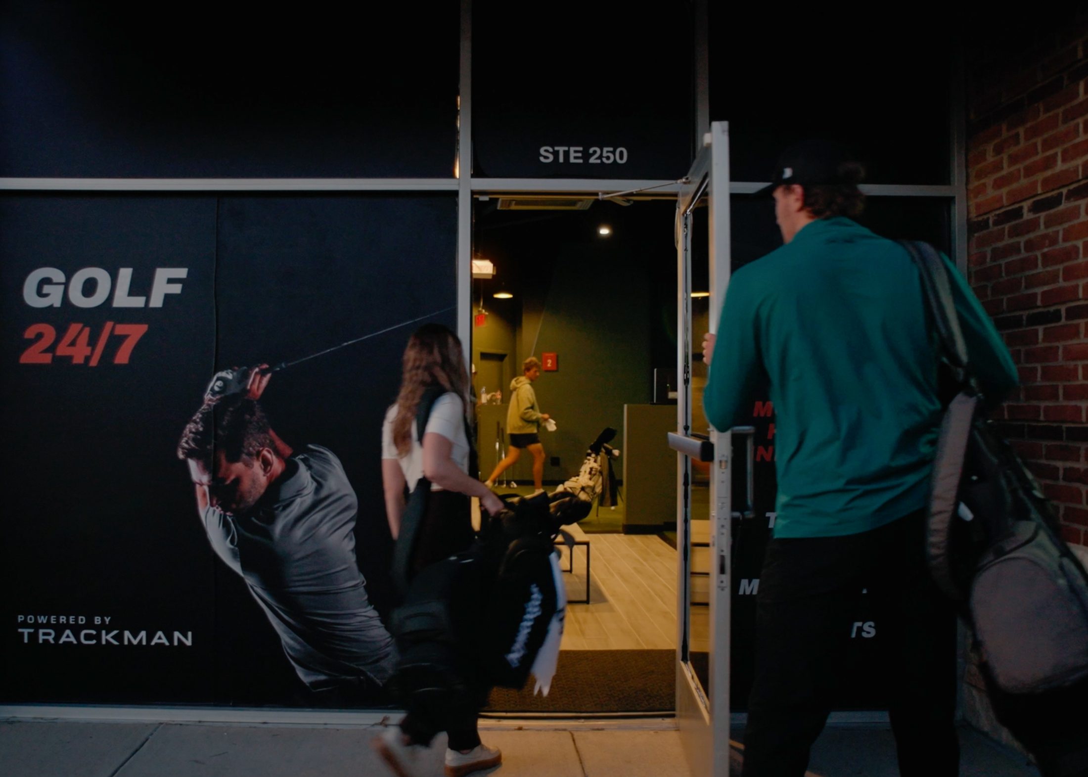 The Tour Studio Golf storefront at night, a window graphic reading GOLF 24/7 powered by Trackman, with members carrying bags through the open door.
