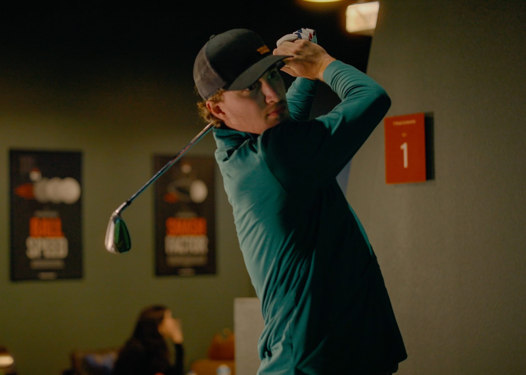 Close portrait of a golfer at the top of his follow-through inside the studio, bay number one and Trackman speed posters visible behind him.